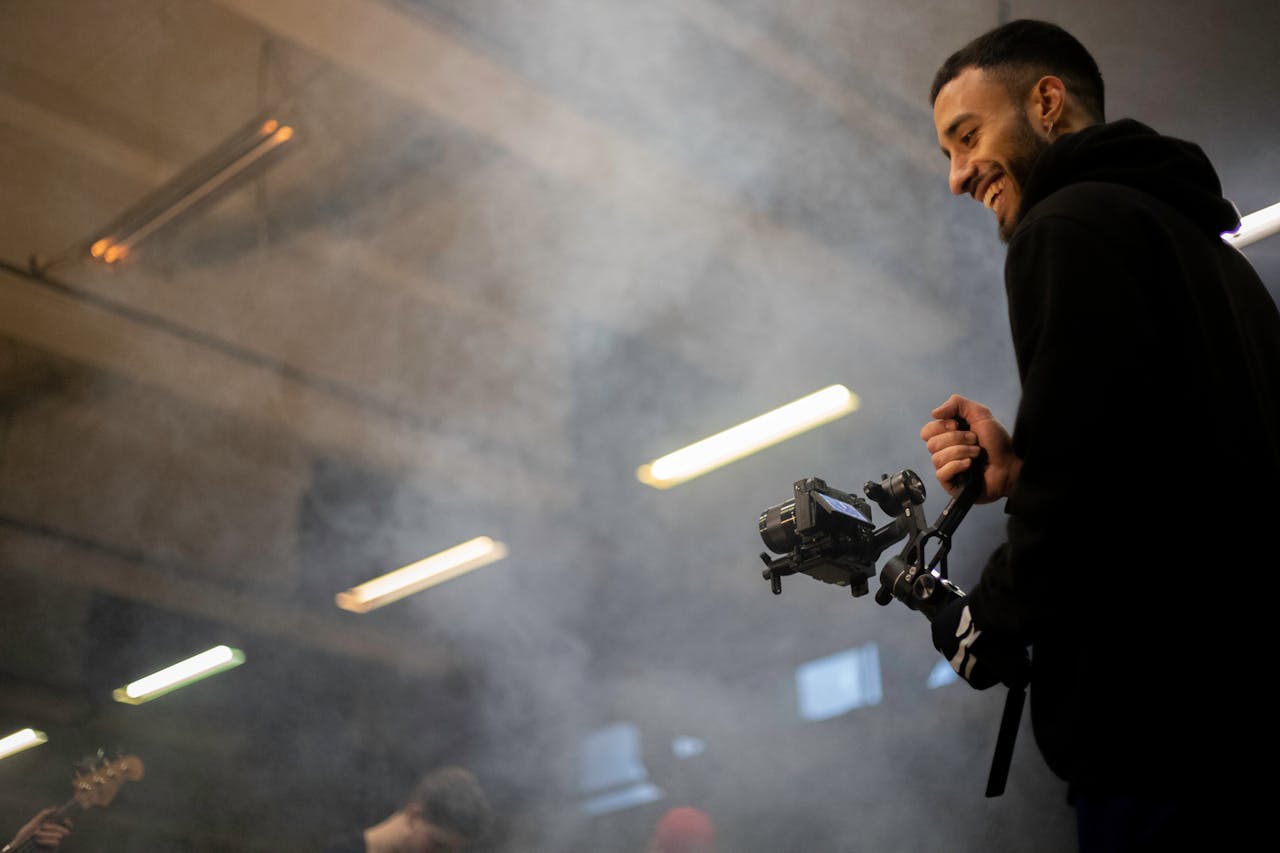 A smiling videographer with equipment amidst a smoky atmospheric indoor environment.